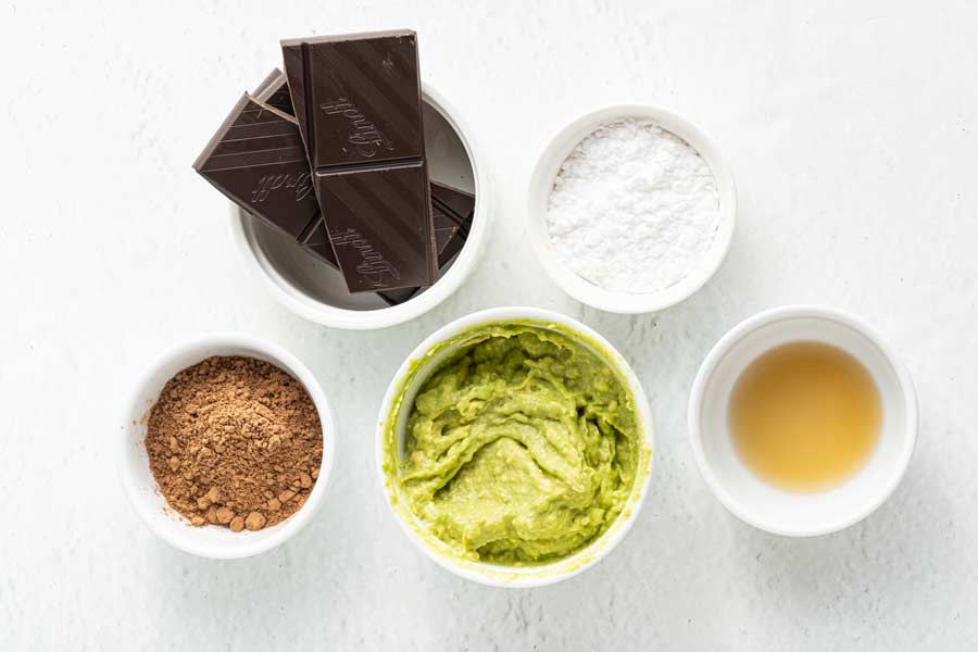 Ingredients for Avocado chocolate keto truffles laid out in separate white bowls on a white surface.
