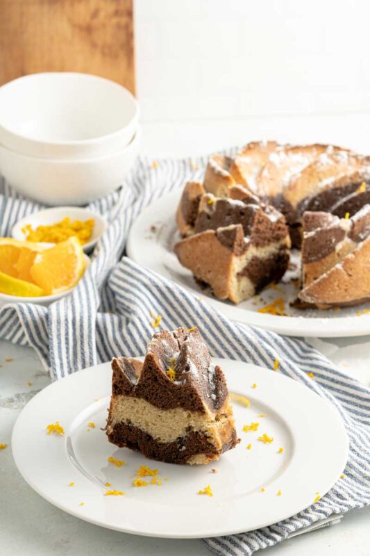 chocolate and orange cake slice placed on a white plate with the whole cake on a plate in the back ground.
