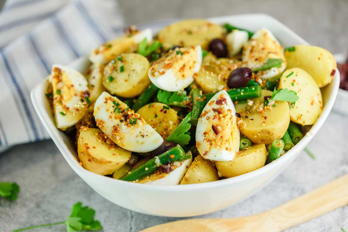 A white bowl filled with a mixed salad containing halved boiled potatoes, quartered hard-boiled eggs, green beans, olives, and topped with mustard seeds dressing and fresh parsley, placed on a table with a wooden spoon nearby.